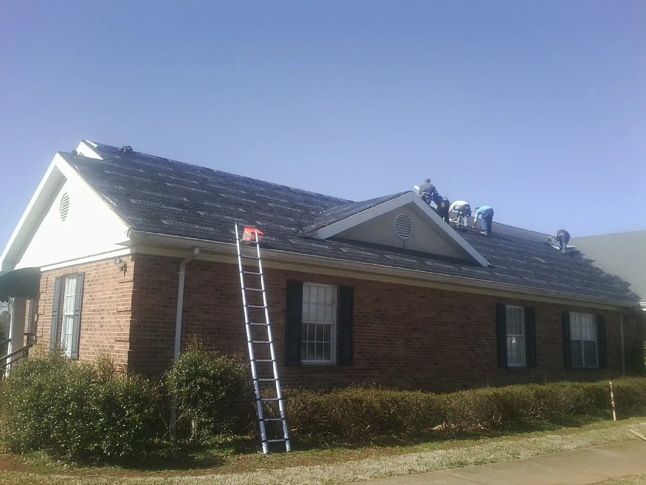 Crew tearing shingles off a brick ranch roof with a ladder set — Metal Roof Repair work by our crew in Isle