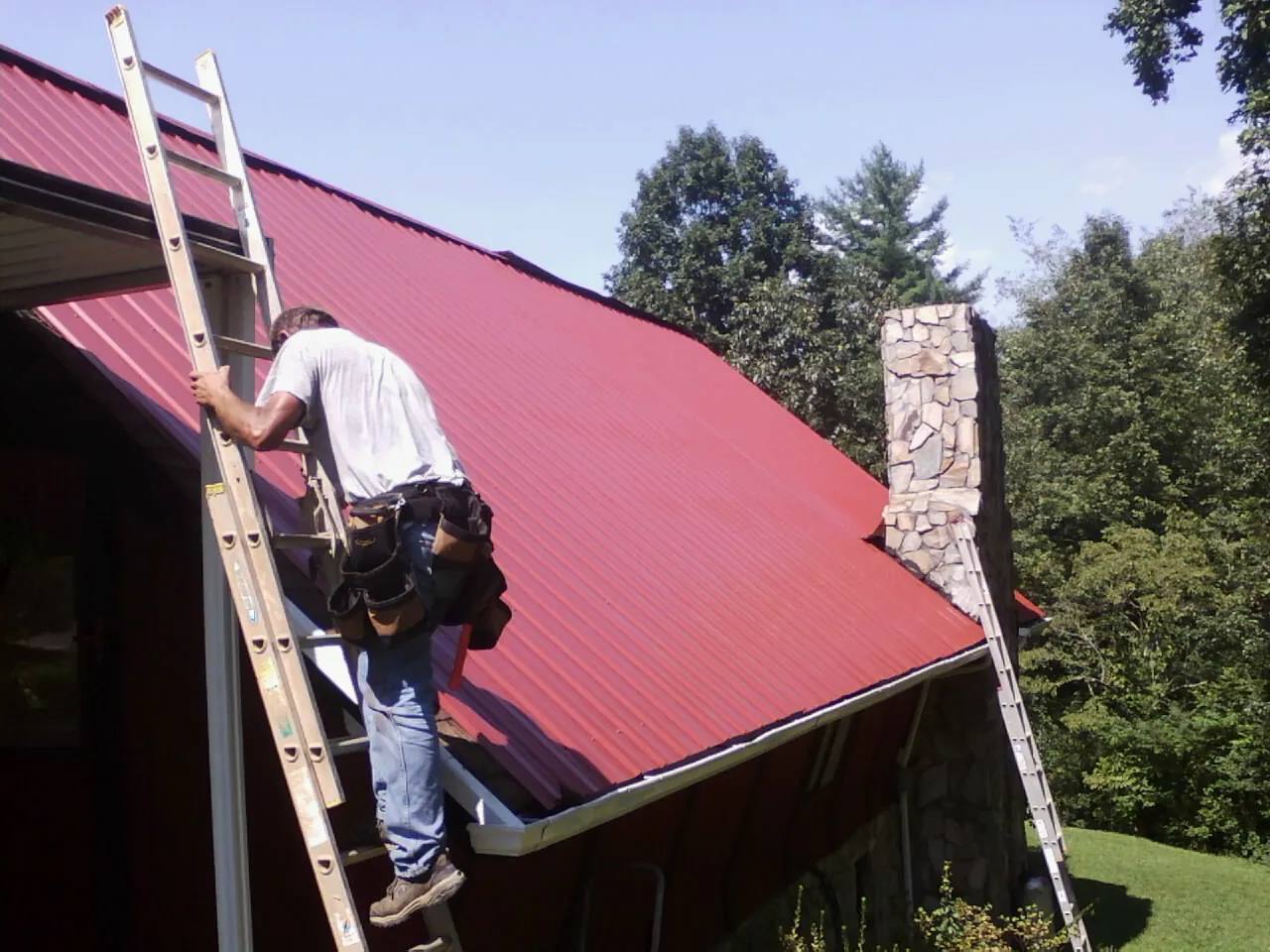 Roofer on a ladder beside a red metal roof and stone chimney — Soffit & Fascia Repair by Roofing Brothers in Isle